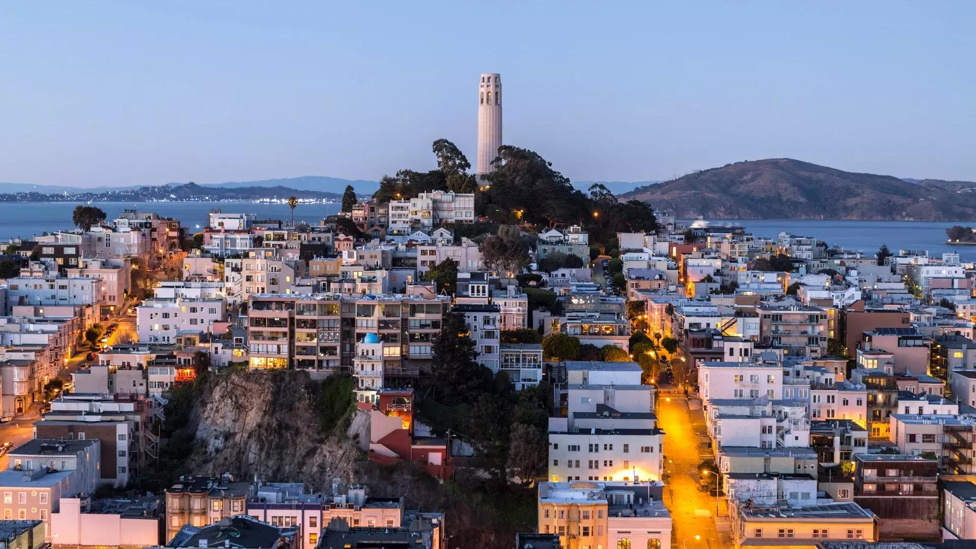 La Coit Tower de San Francisco au crépuscule, avec des rues éclairées devant elle et la baie de San Francisco derrière elle.