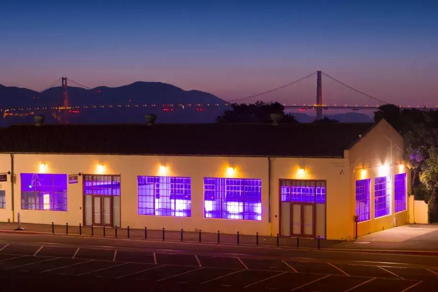 O edifício Fort Mason é iluminado por uma luz interna roxa à noite, com a ponte ao fundo.